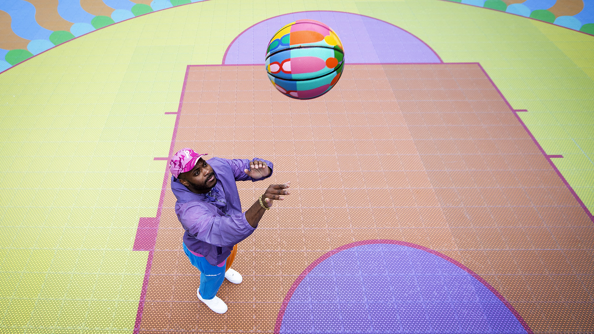 Man in a purple jacket and blue trousers looking up at a brightly coloured basketball in the air on a coloured court 