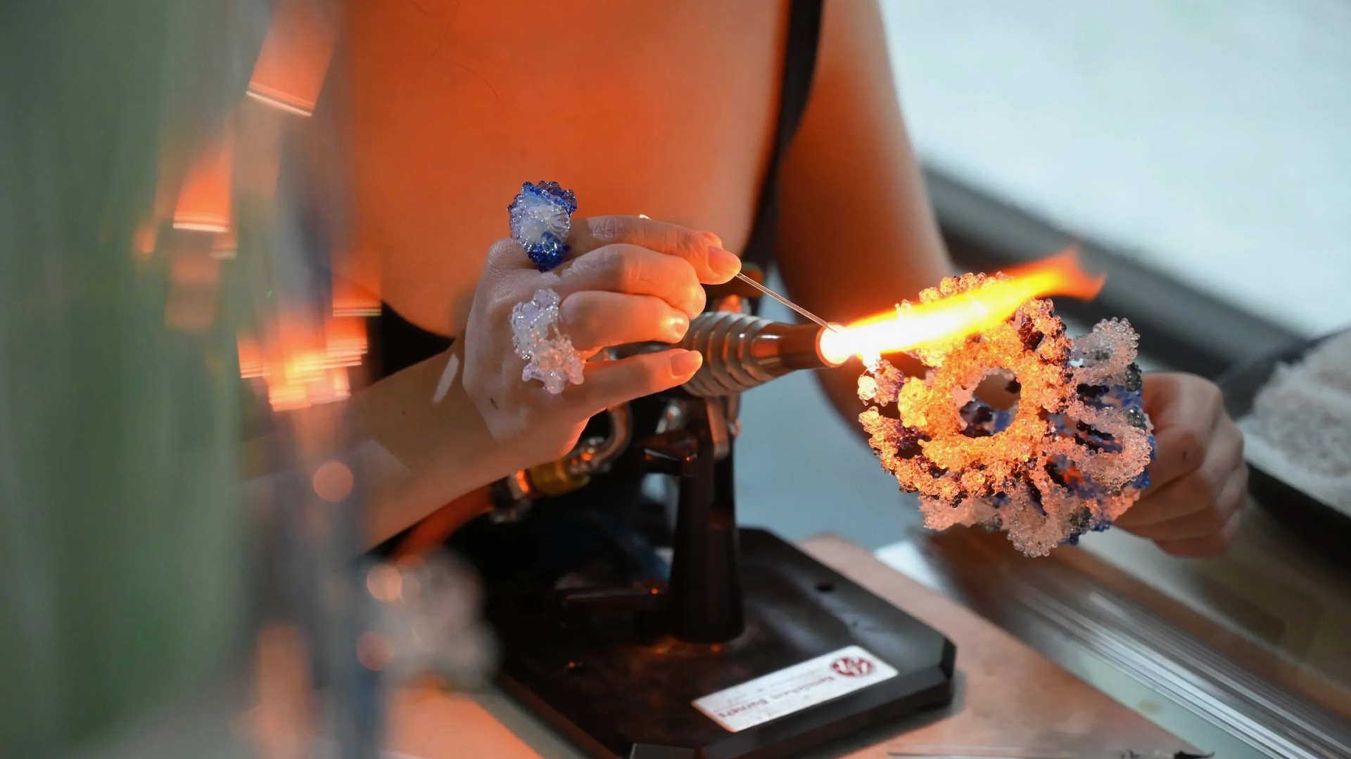 Close-up of hands using a torch flame to shape a detailed blue and clear glass flower.