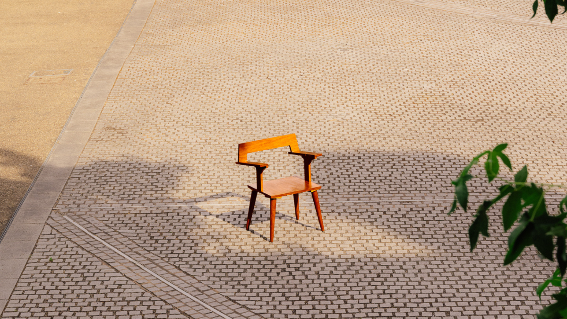 A handcrafted wooden chair sits in a sunlit cobbled courtyard with long tree shadows across the paving.