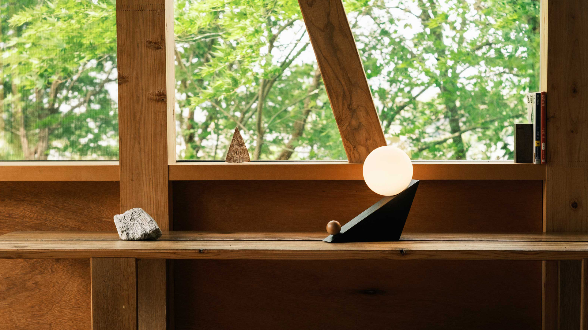 TILT portable lamp glowing on a wooden shelf beside large windows overlooking green trees.
