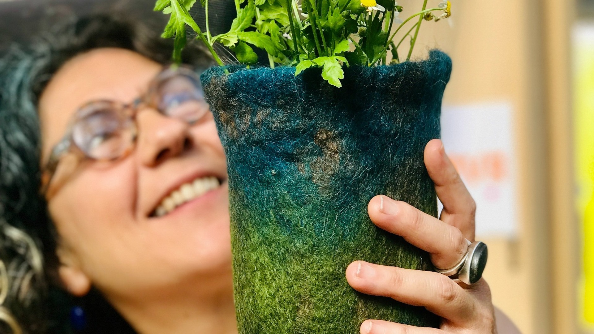 Designer of MINOVA smiling while holding a handmade blue-green wool felt vase filled with fresh white flowers