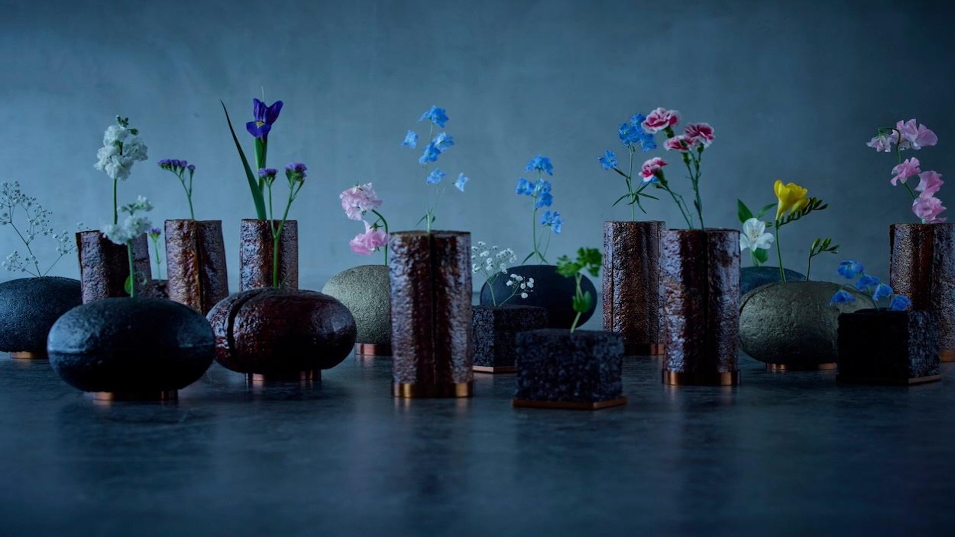 Natural material vessels displayed with colourful flowers in a dark blue setting, creating a sculptural garden-like arrangeme