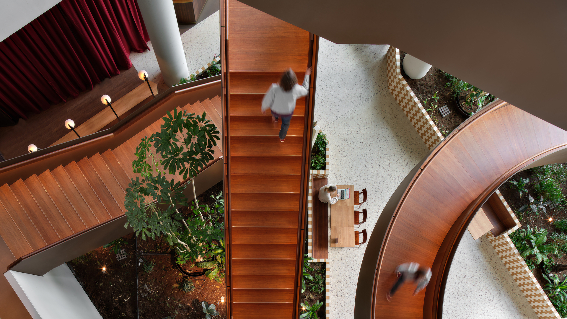 Top view of timber staircases at The Pulse Amsterdam, with people moving between levels and lush green indoor planting below.