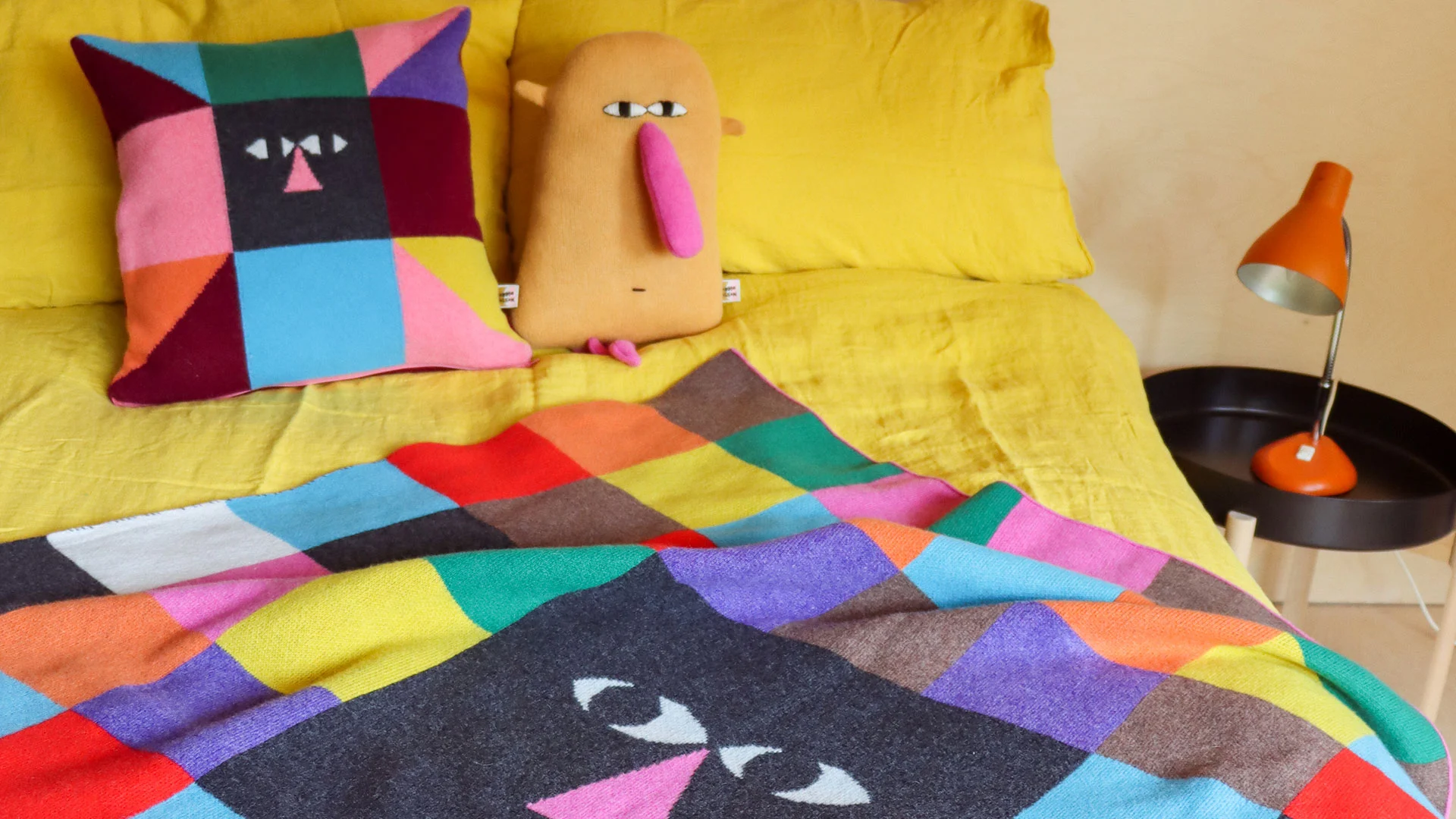 A bold, multicolour knitted cushion and throw draped across a bed, with a knitted yellow character sitting alongside them.