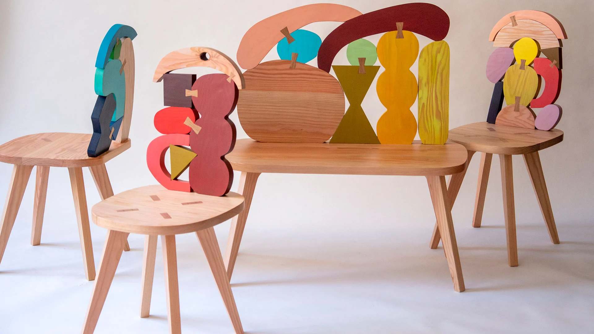 Three wooden sculptural chairs and a bench, painted in a colourful palette.