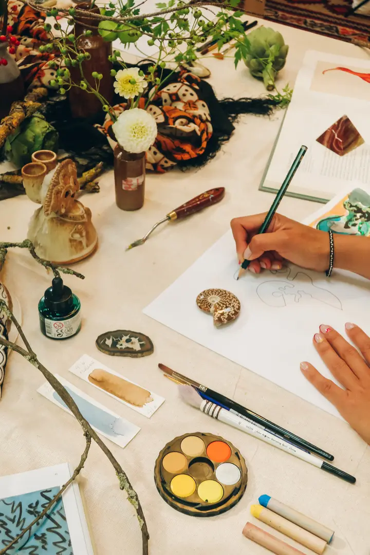 close-up of artist sketching on paper with miscellaneous items surrounding the table