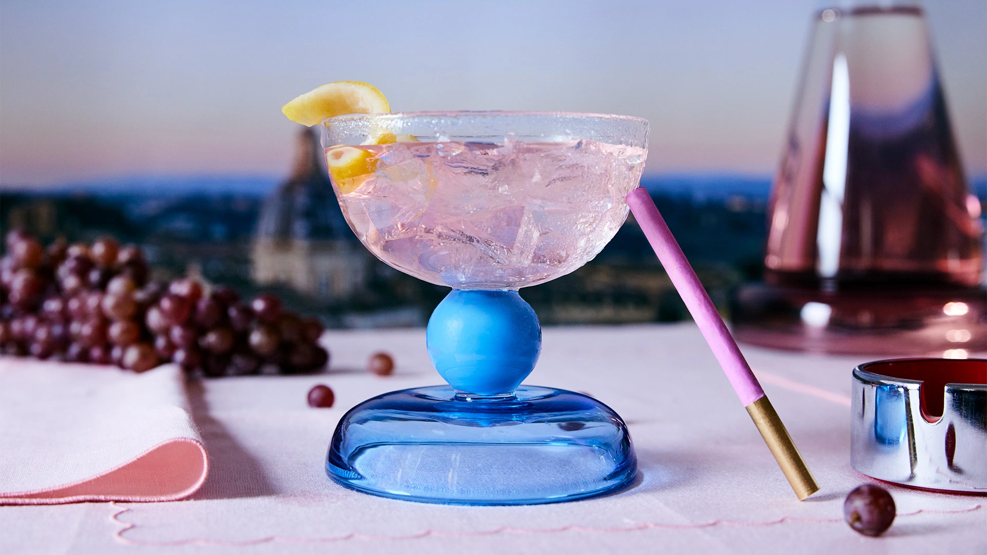 Blue cocktail glass filled with liquid in foreground, with aubergine colour carafe in background