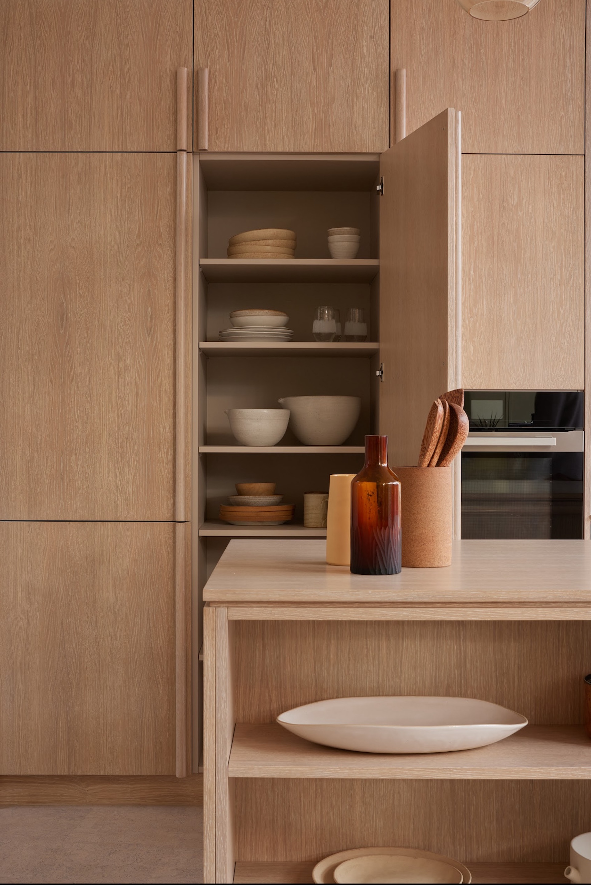 A close-up view of a minimalist kitchen with light wood cabinetry. One tall cabinet door is open, revealing neatly arranged s