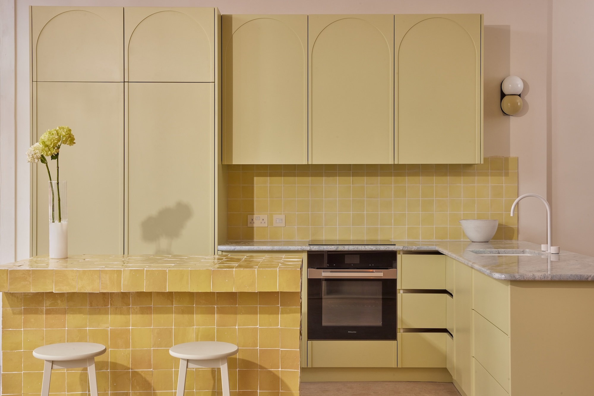 A contemporary kitchen with a soft yellow color palette. The cabinetry features flat panel doors with arched detailing on the