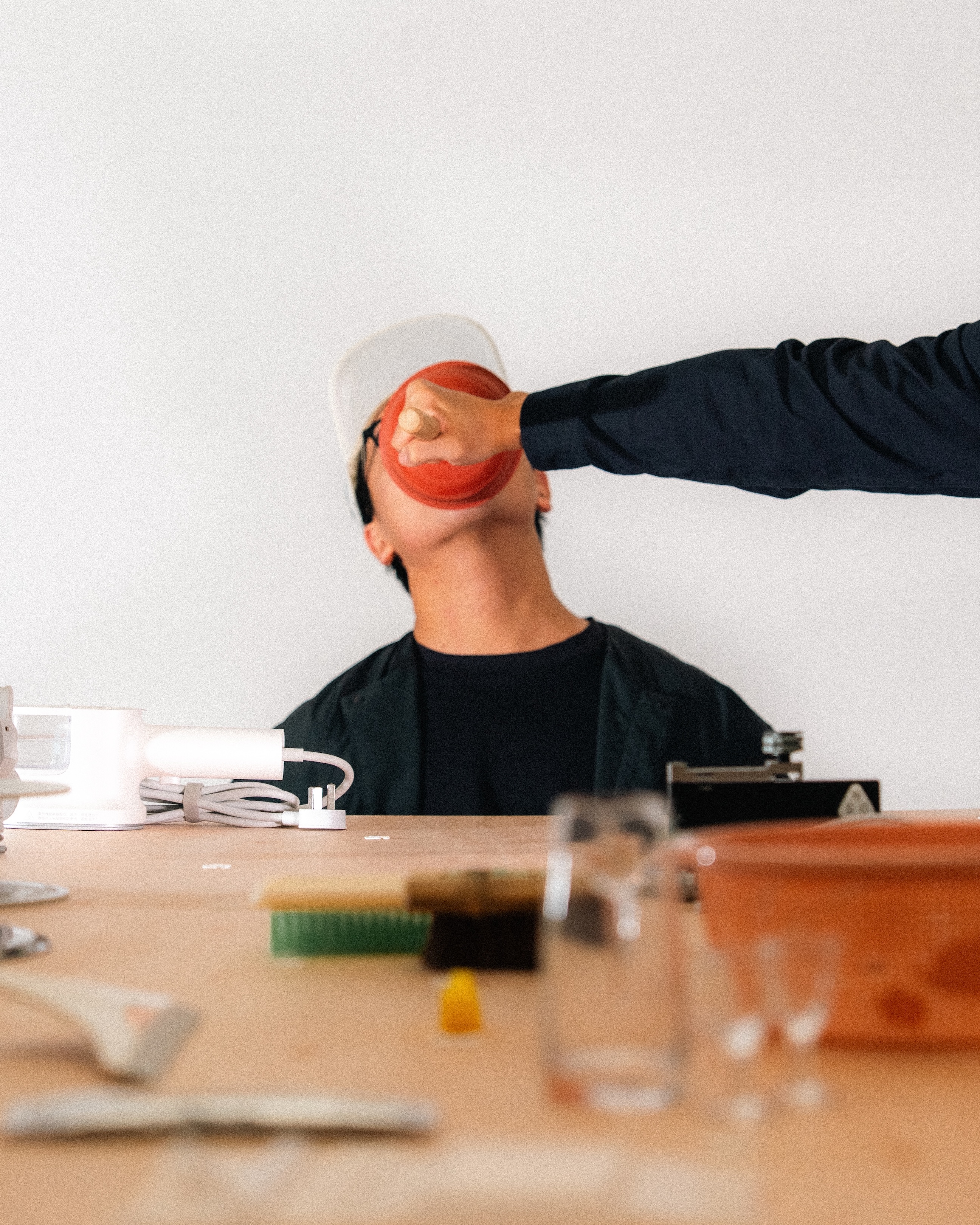 Person wearing a cap leans back as another hand presses a red rubber plunger against their face.
