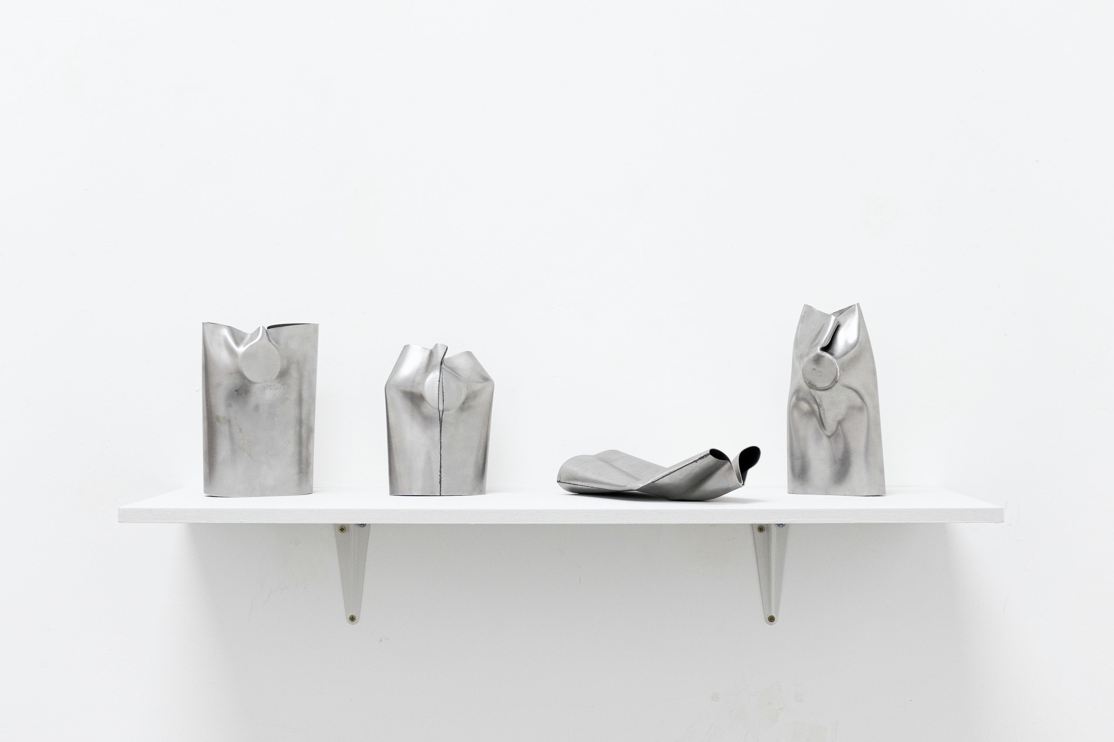 Four sculptural crushed metal vessels displayed on a white wall-mounted shelf, minimalist and industrial in style.