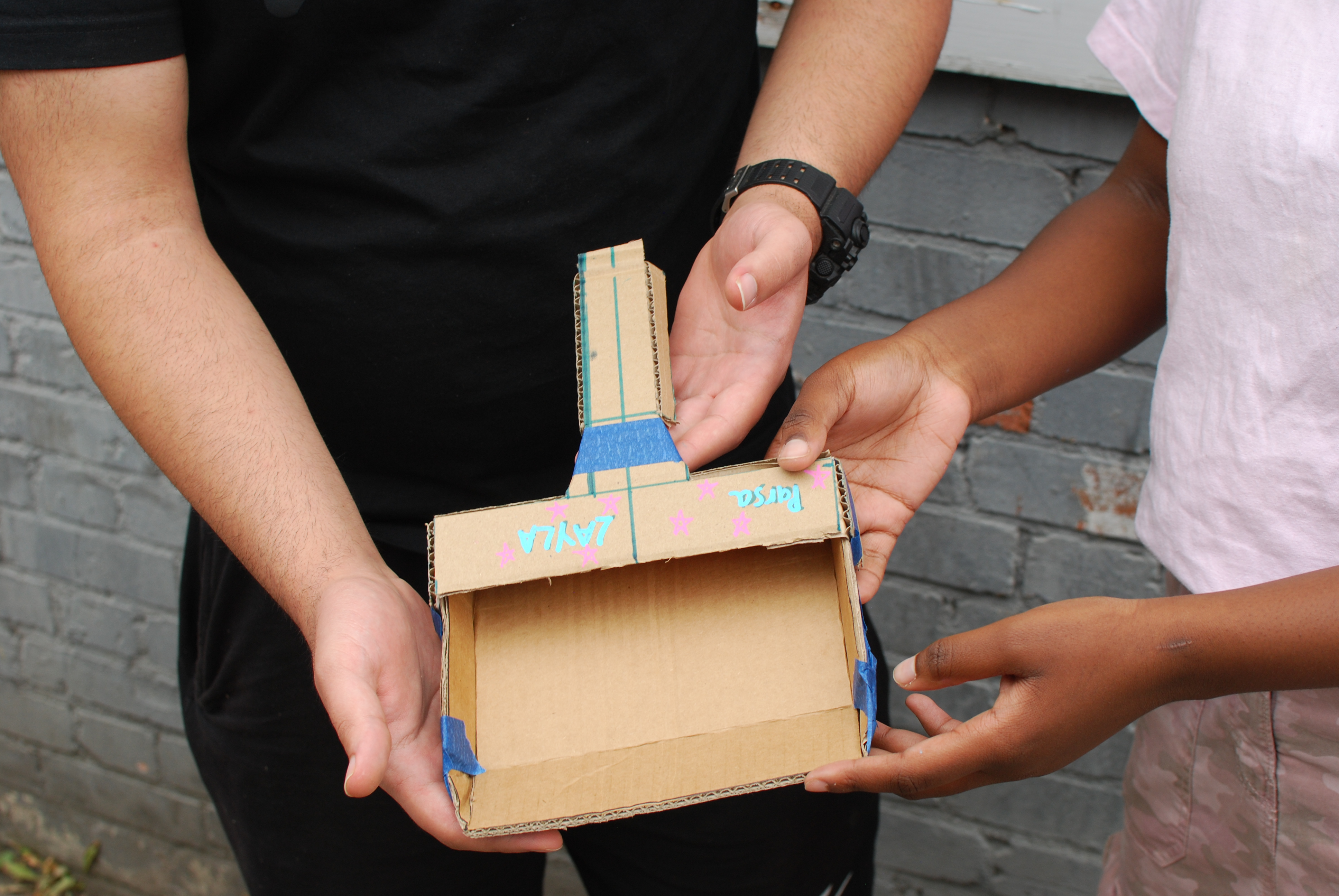 Two young people hold up a model dustpan made of cardboard, with coloured writing on it, in front of a grey brick wall.