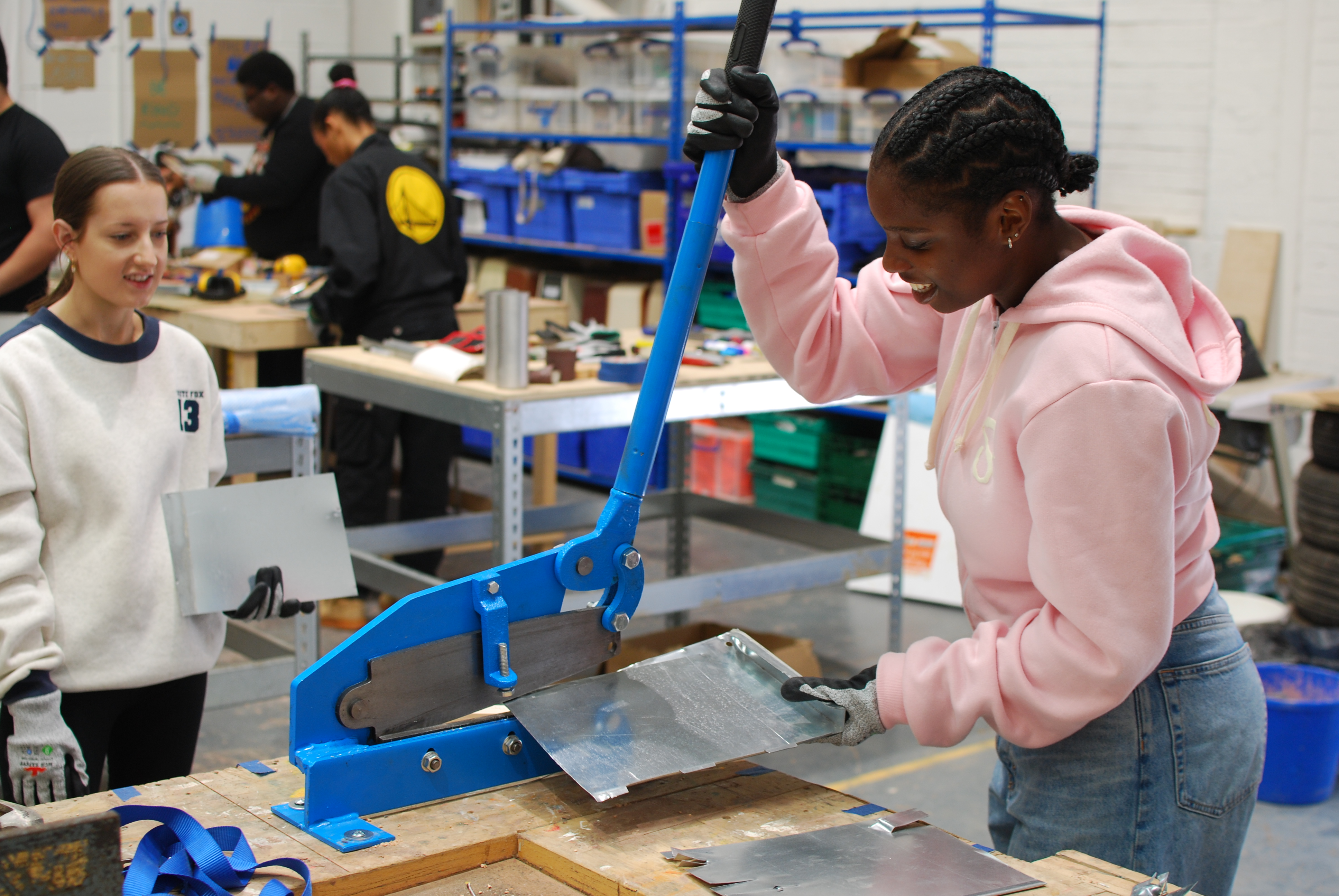 A young woman in a pink hoodie uses a blue metal guillotine to cut scrap metal.