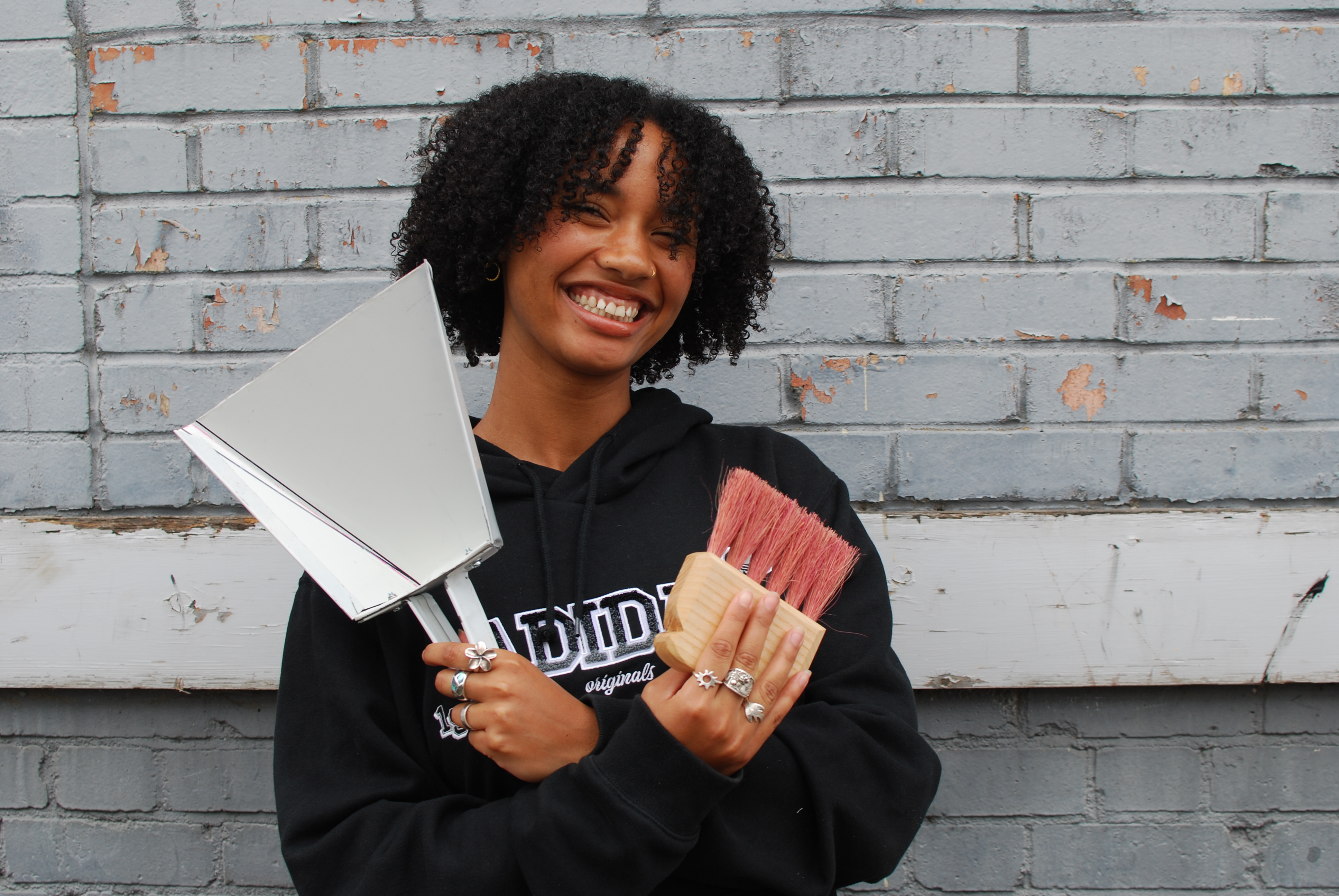 A young person holds up a prototype dustpan and brush. She is wearing a black hoodie and has her arms crossed. 