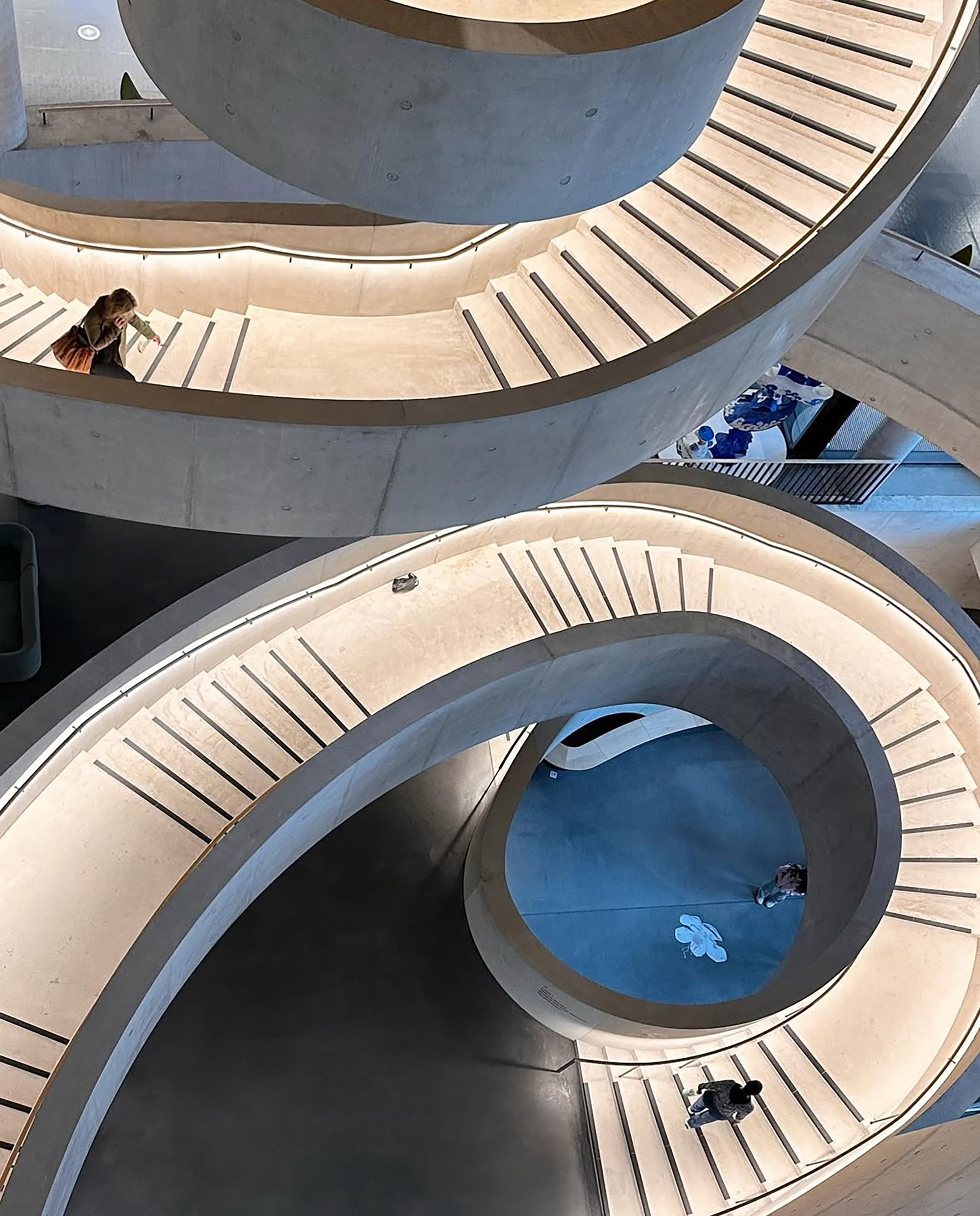 A image of a light concrete spiral staircase from above that is illuminated by warm light with people walking on it