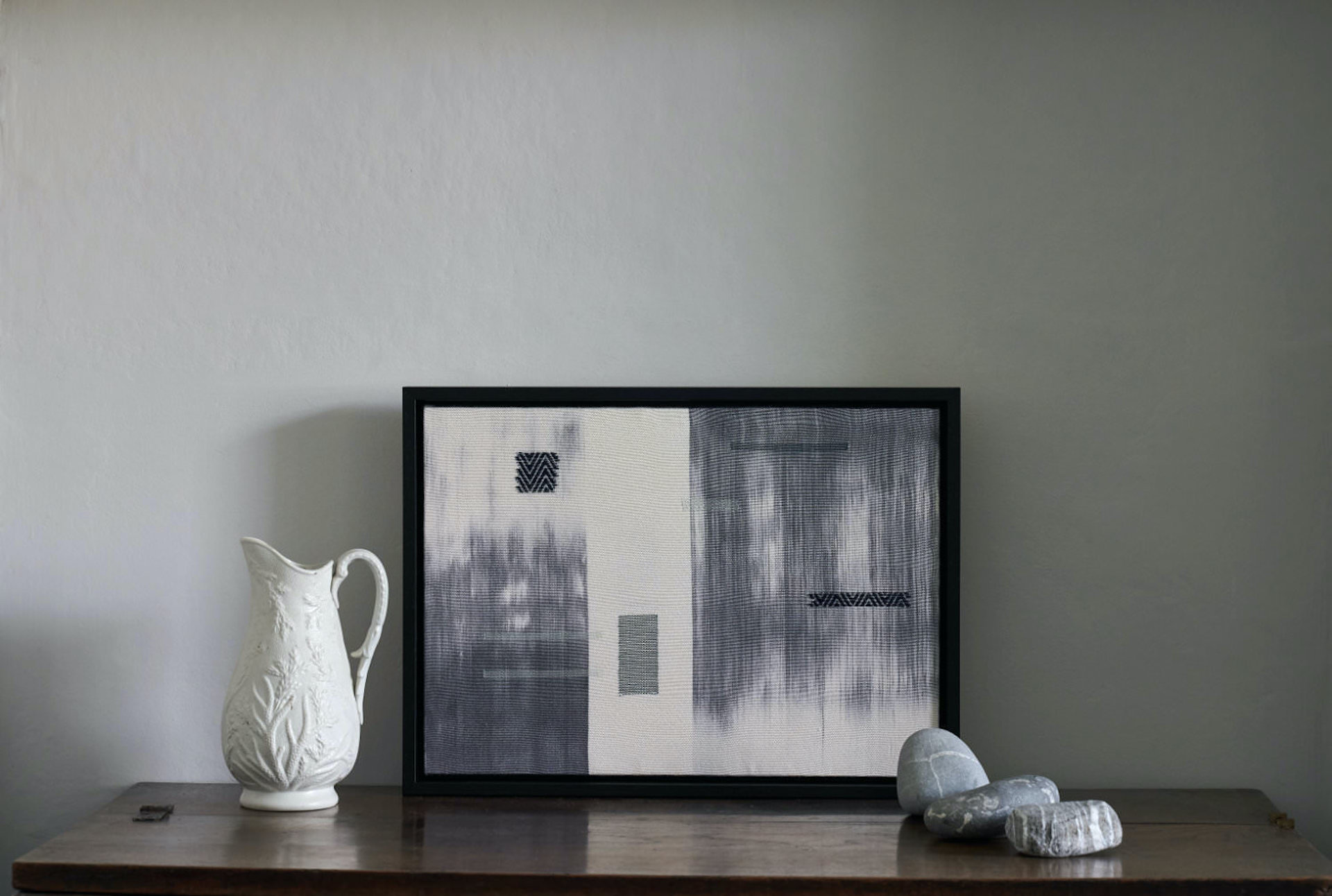A framed woven artwork in tones of white, blue and grey leaning against a wall on top of a dresser